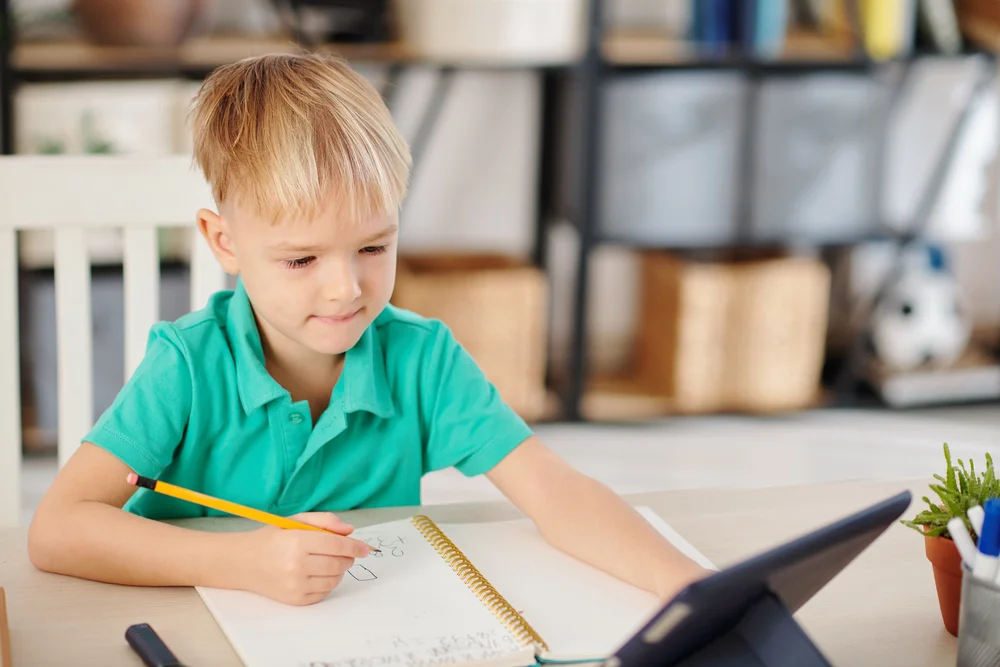 Young boy writing in notebook at desk using pencil, online learning