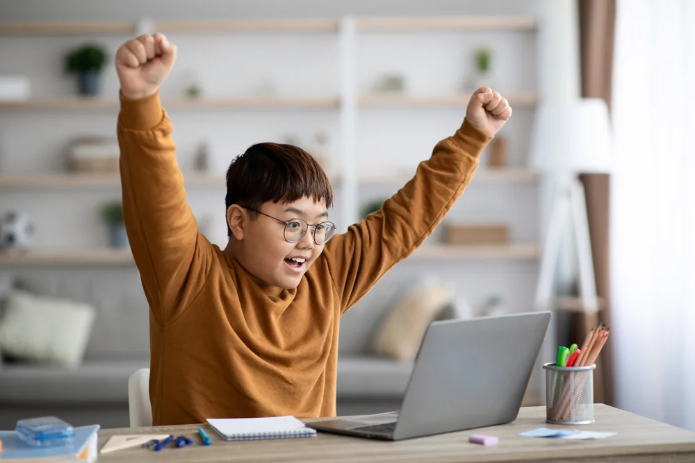 Excited boy celebrates online learning success, arms raised in victory.