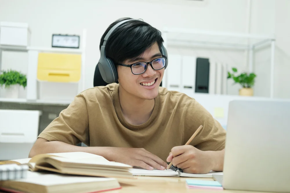 Smiling Asian man wearing headphones studies online at his desk, taking notes.