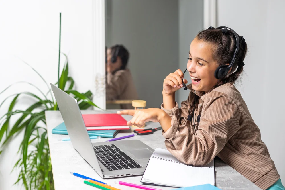 Happy girl in headset participates in online class, using laptop and notebook.