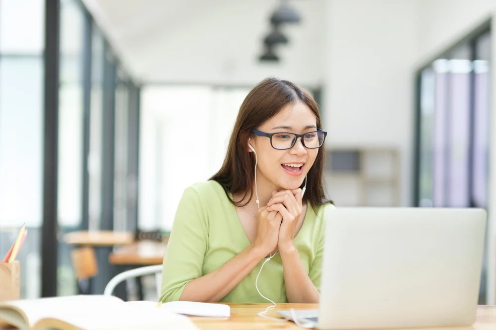 Smiling woman using laptop for online video call, wearing glasses and earphones.