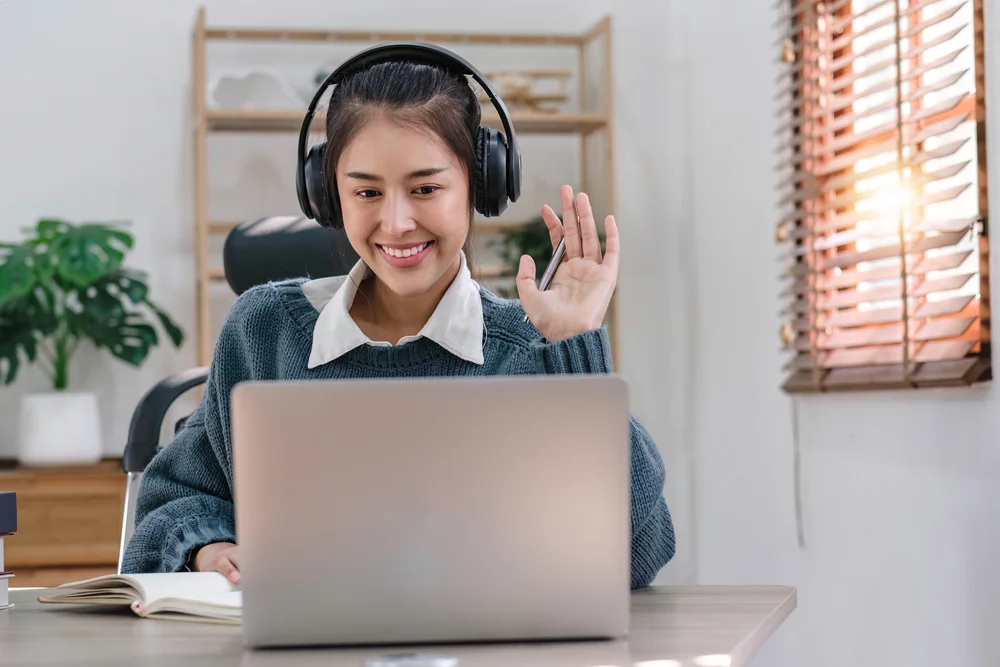 Woman on video call, waving and smiling while working from home on laptop.