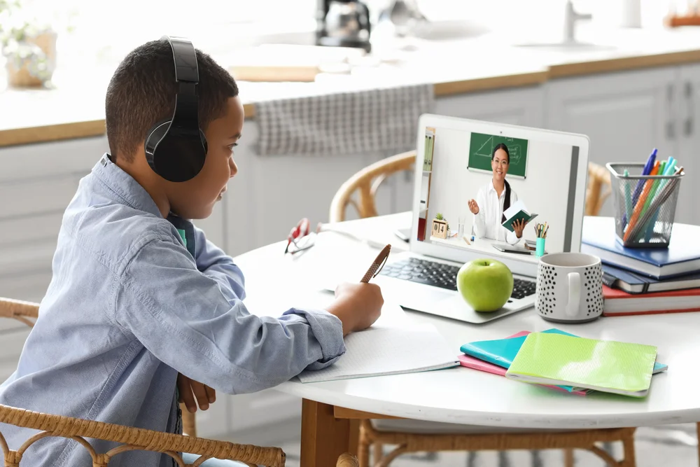 Boy wearing headphones participates in online class via laptop, taking notes.